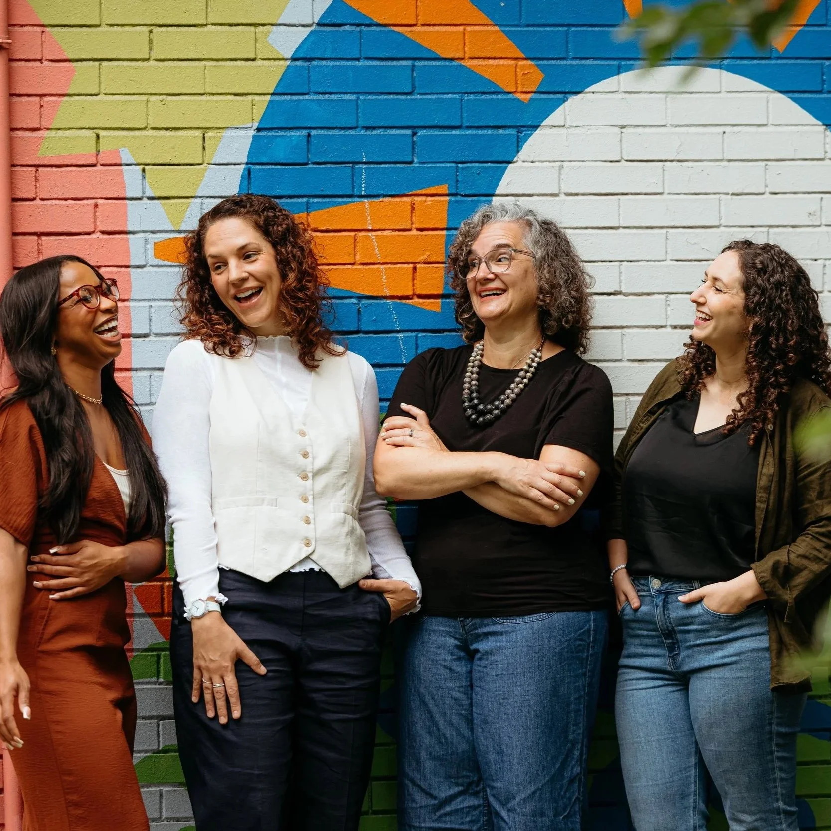 Group of four women laughing together in front of a colorful mural painted on a brick wall.