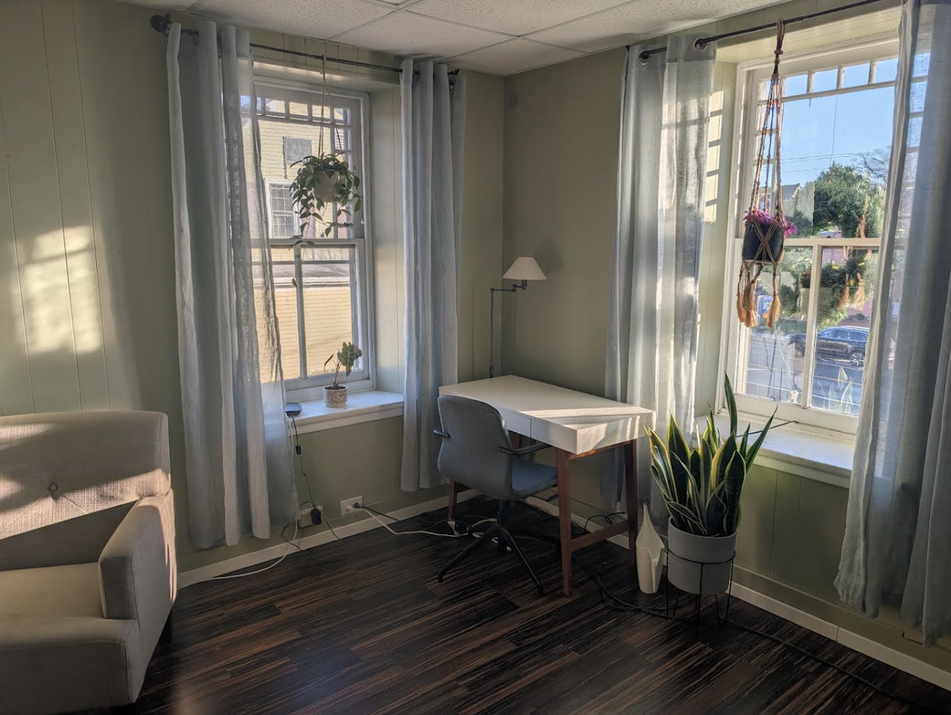 A corner of a room with light green walls, white curtains, and large windows. There is a white armchair partially visible on the left, a white desk with a gray office chair, two potted plants, and a standing lamp.