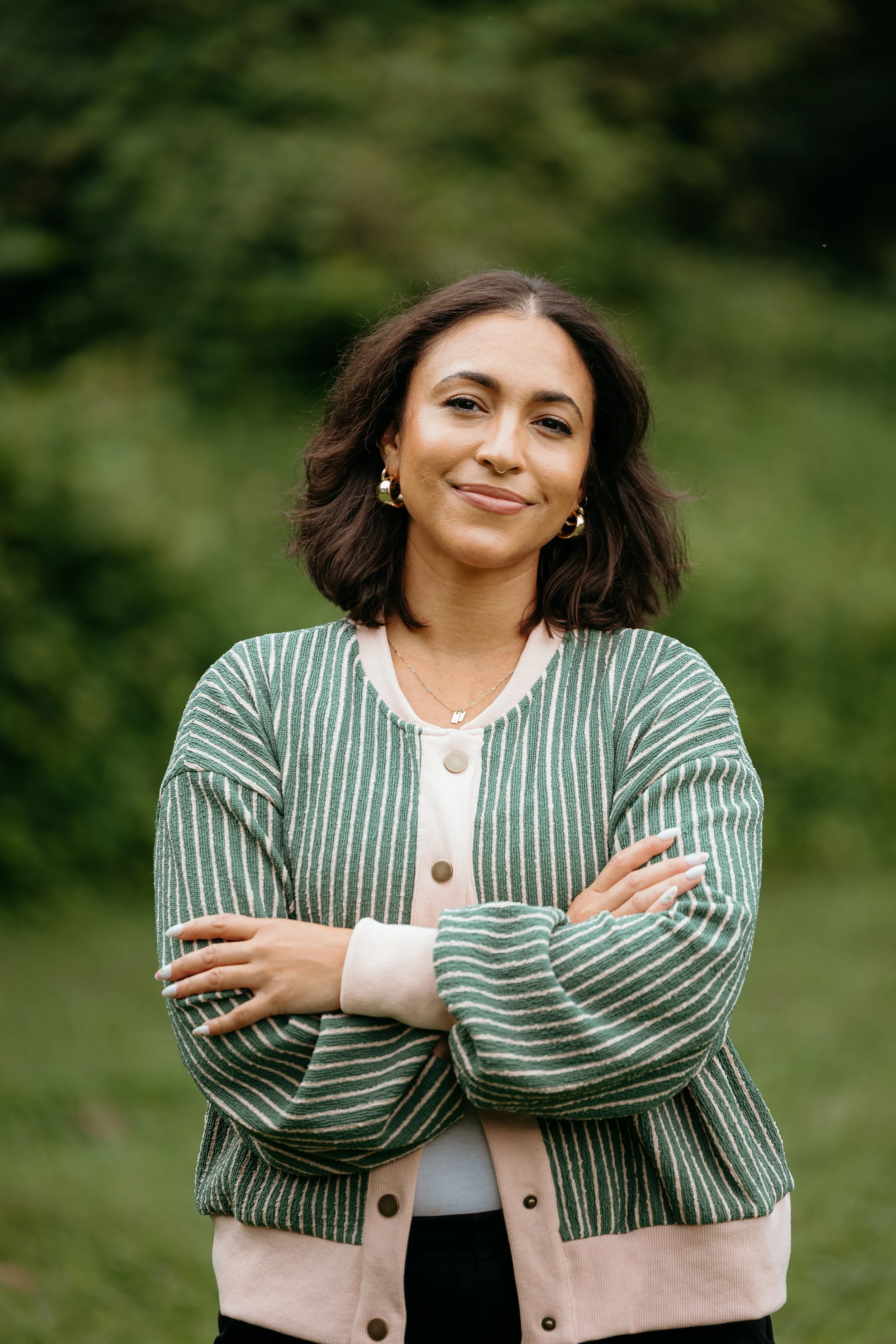 A woman with shoulder-length dark brown hair, wearing a striped green and beige cardigan, standing outdoors with her arms crossed and smiling softly at the camera.