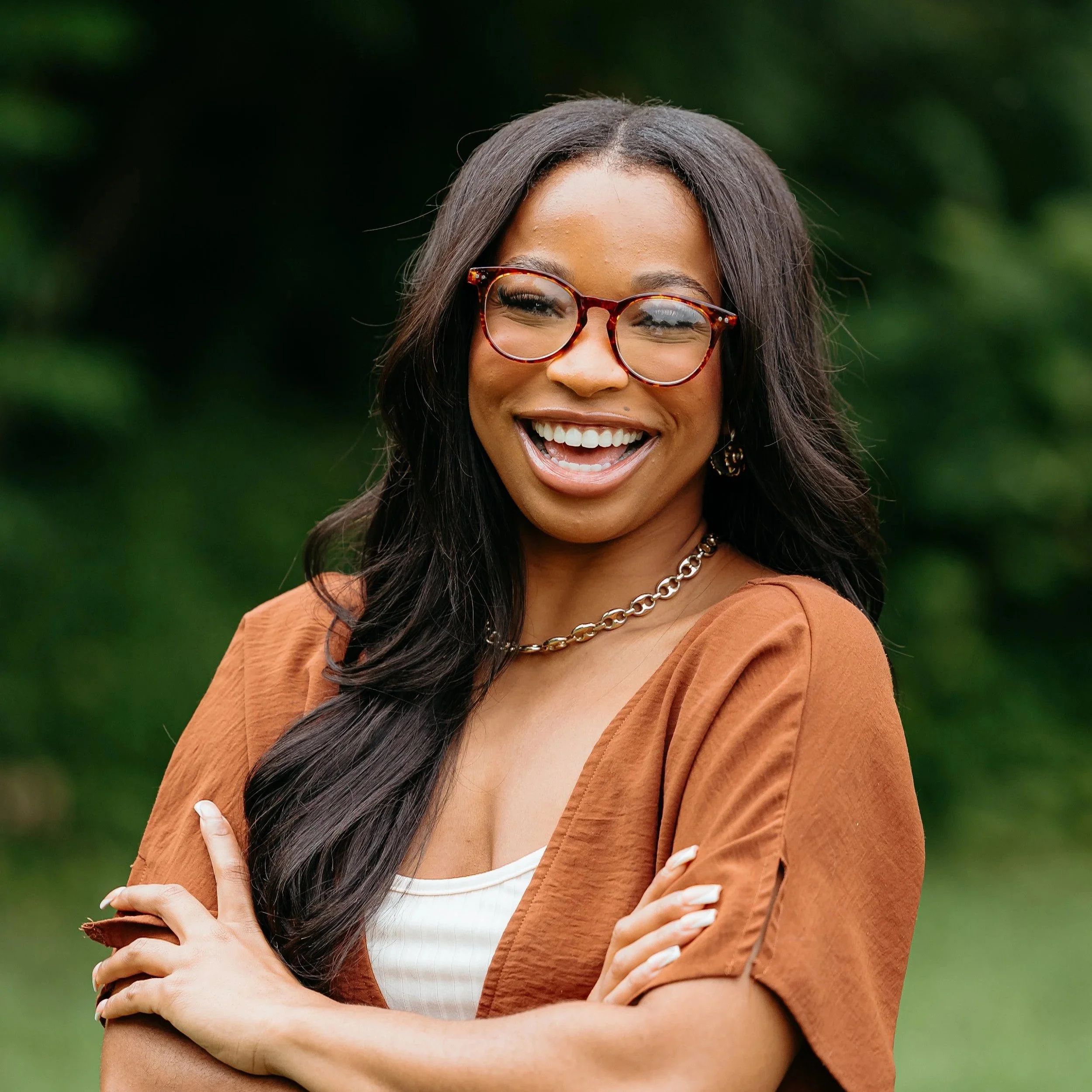 A woman with long dark hair, wearing tortoiseshell glasses, smiling and crossing her arms, dressed in a tan jacket and white top, outdoors with a green blurred background.