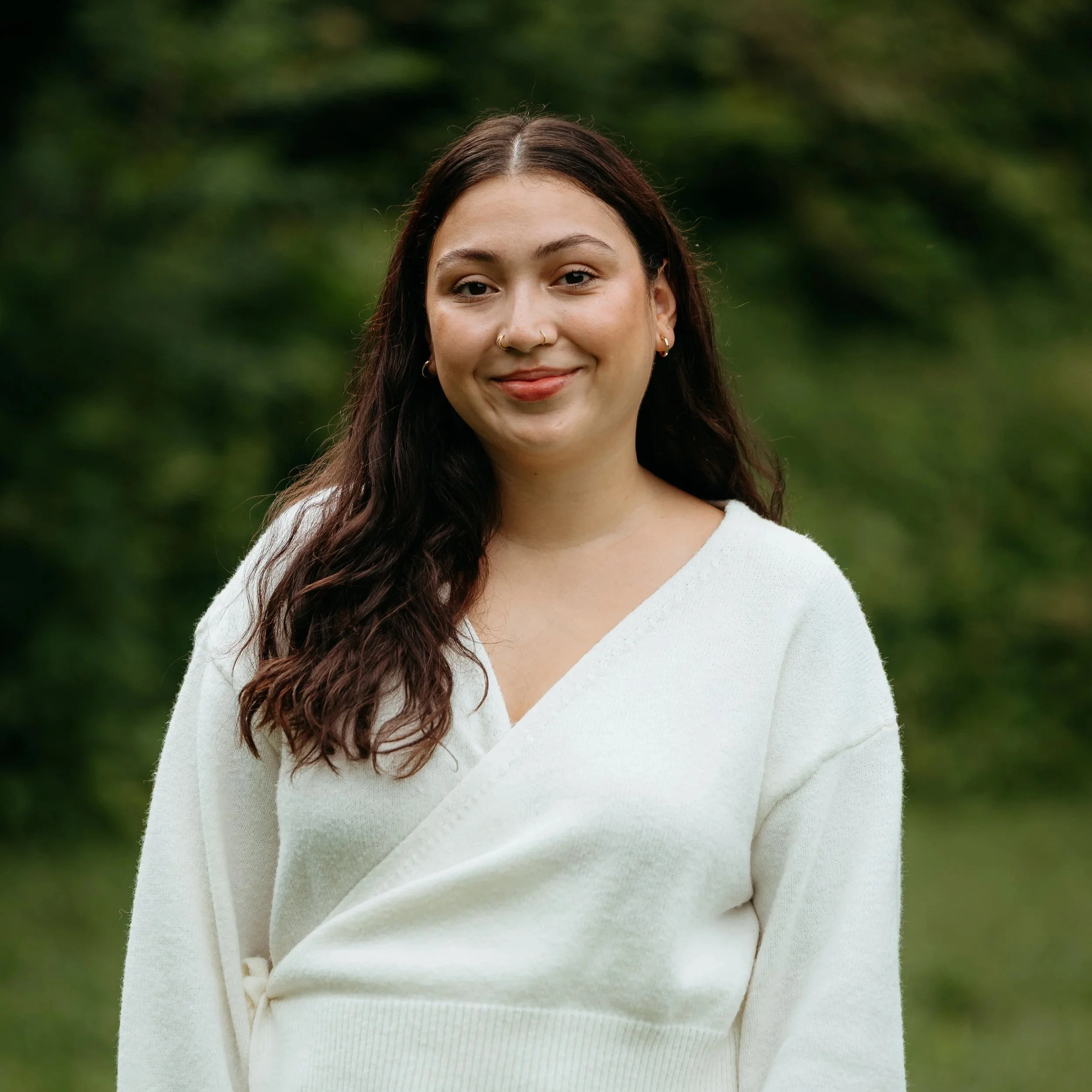 Close-up of a young woman with long dark hair, wearing a white V-neck sweater, standing outdoors against a blurred green background.