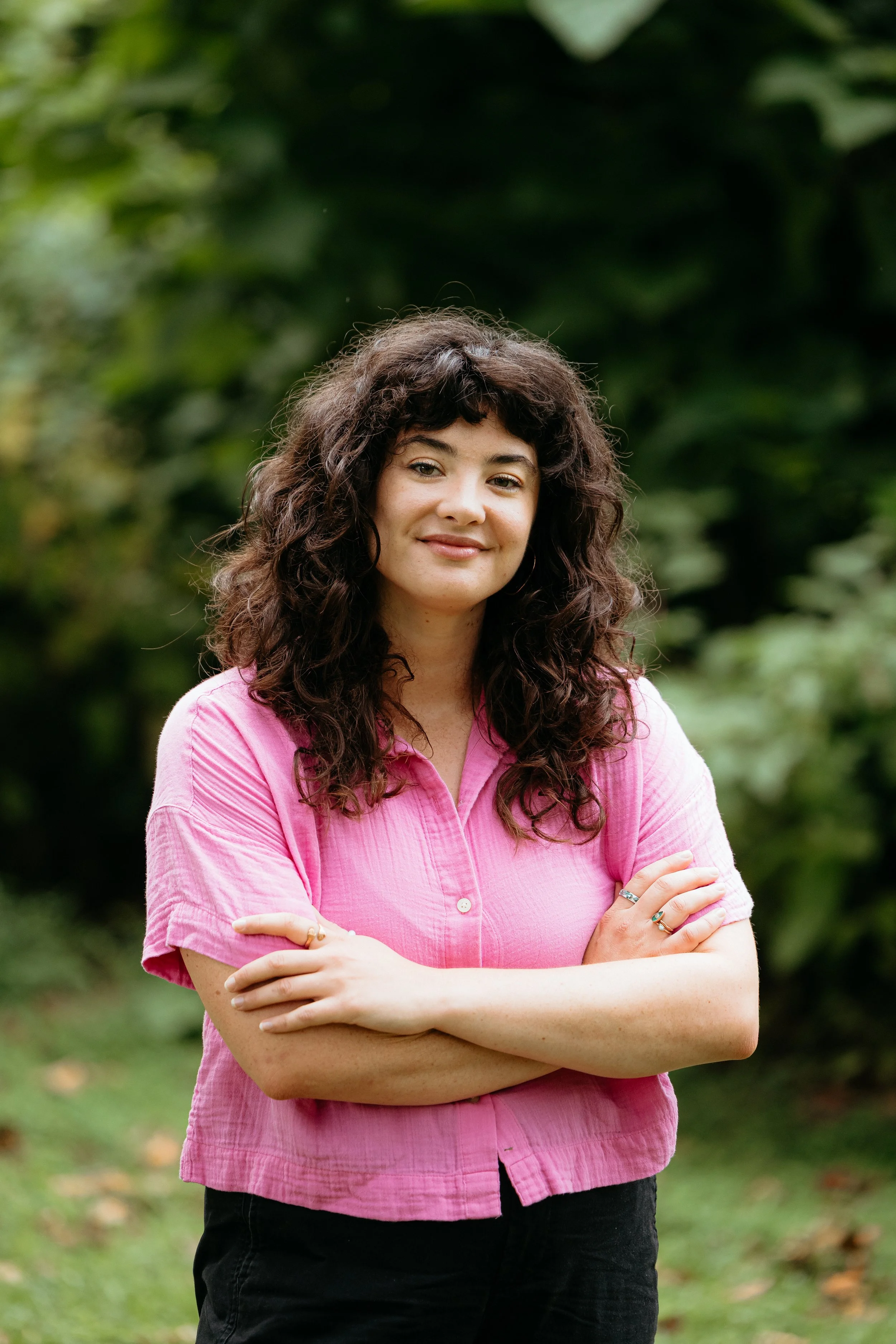 A woman with dark curly hair wearing a pink shirt and black pants, standing outdoors with arms crossed, smiling and looking at the camera, with green foliage in the background.