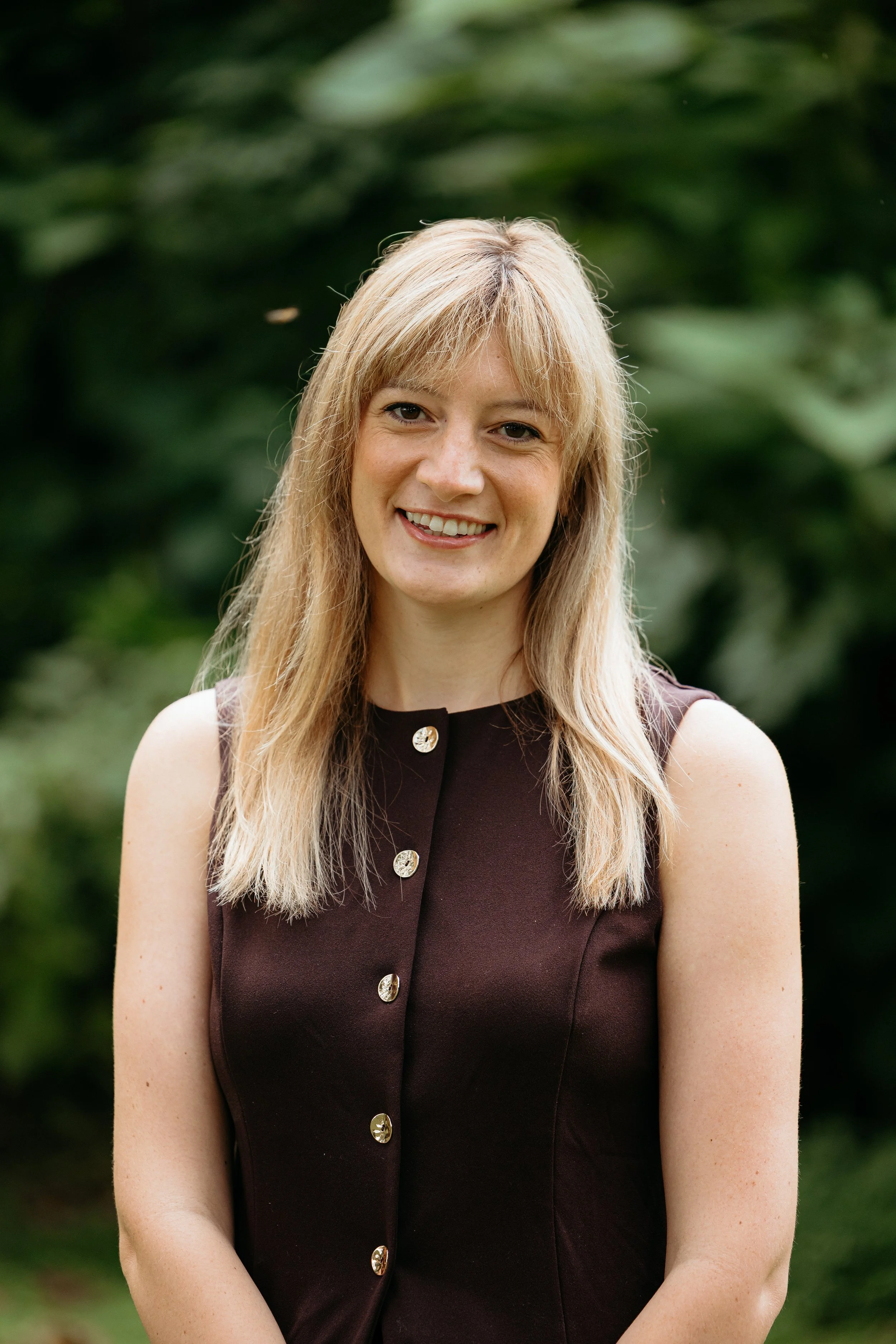 Portrait of a woman with blonde hair wearing a sleeveless dark brown dress with gold buttons, standing outdoors with blurred greenery in the background.