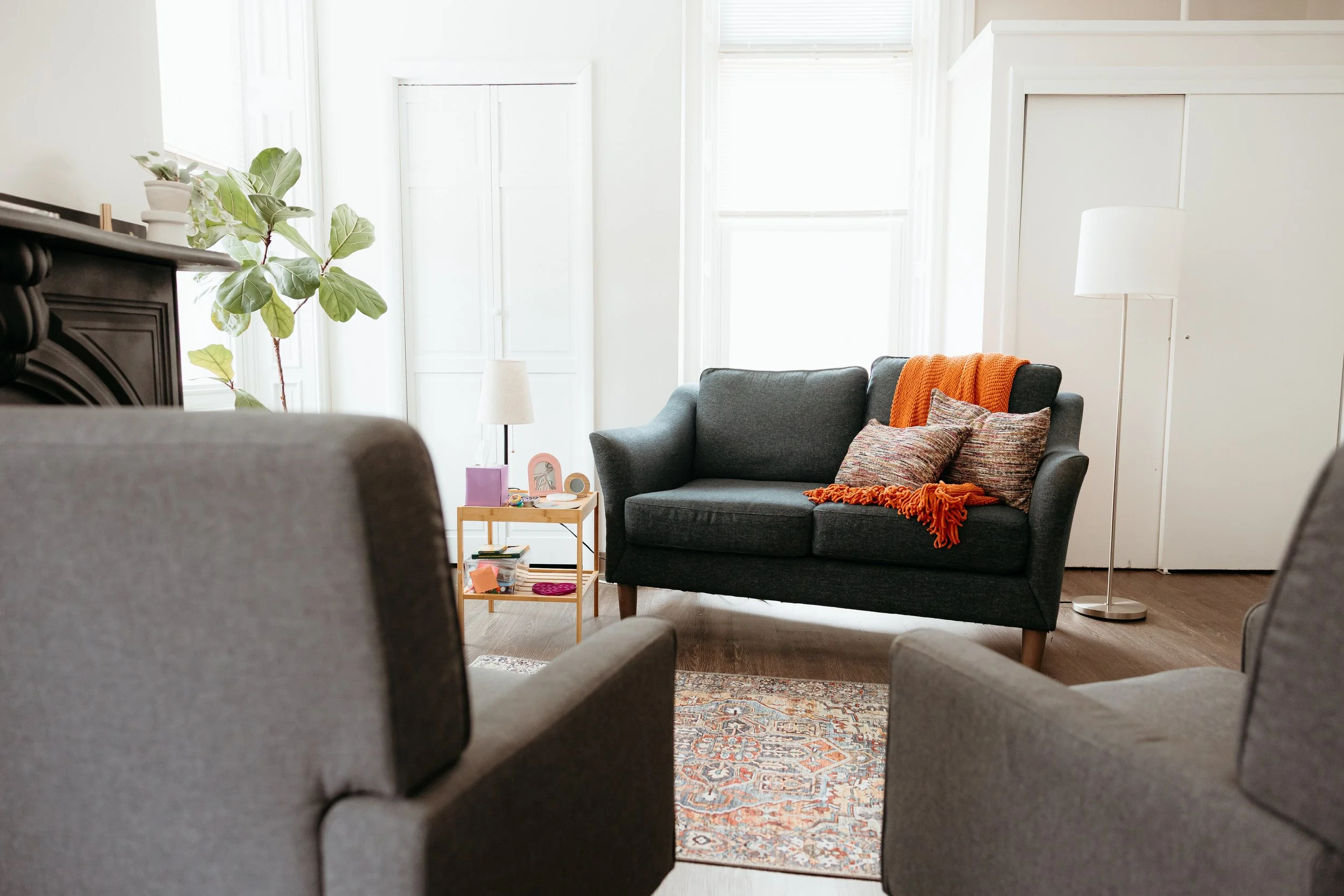 Living room with gray sofa, colorful pillows, orange throw blanket, wooden side table with books, plants, and decorative items, large windows with blinds, white walls, and hardwood floor.