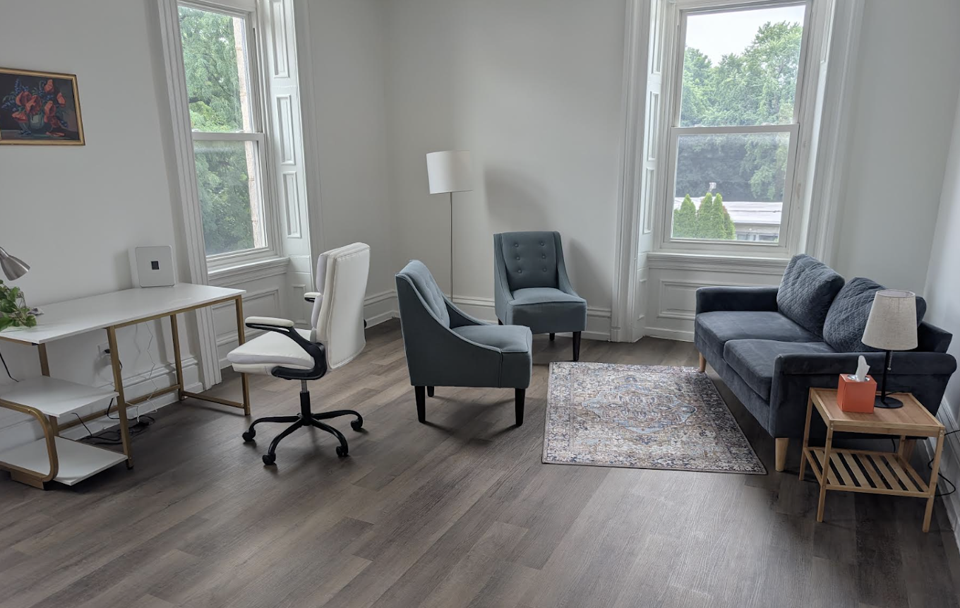 A bright living room with large windows, three blue armchairs, a dark blue sofa, a white desk with a white office chair, a floor lamp, a small wooden side table with a lamp and tissue box, a decorative rug, and a framed picture on the wall.