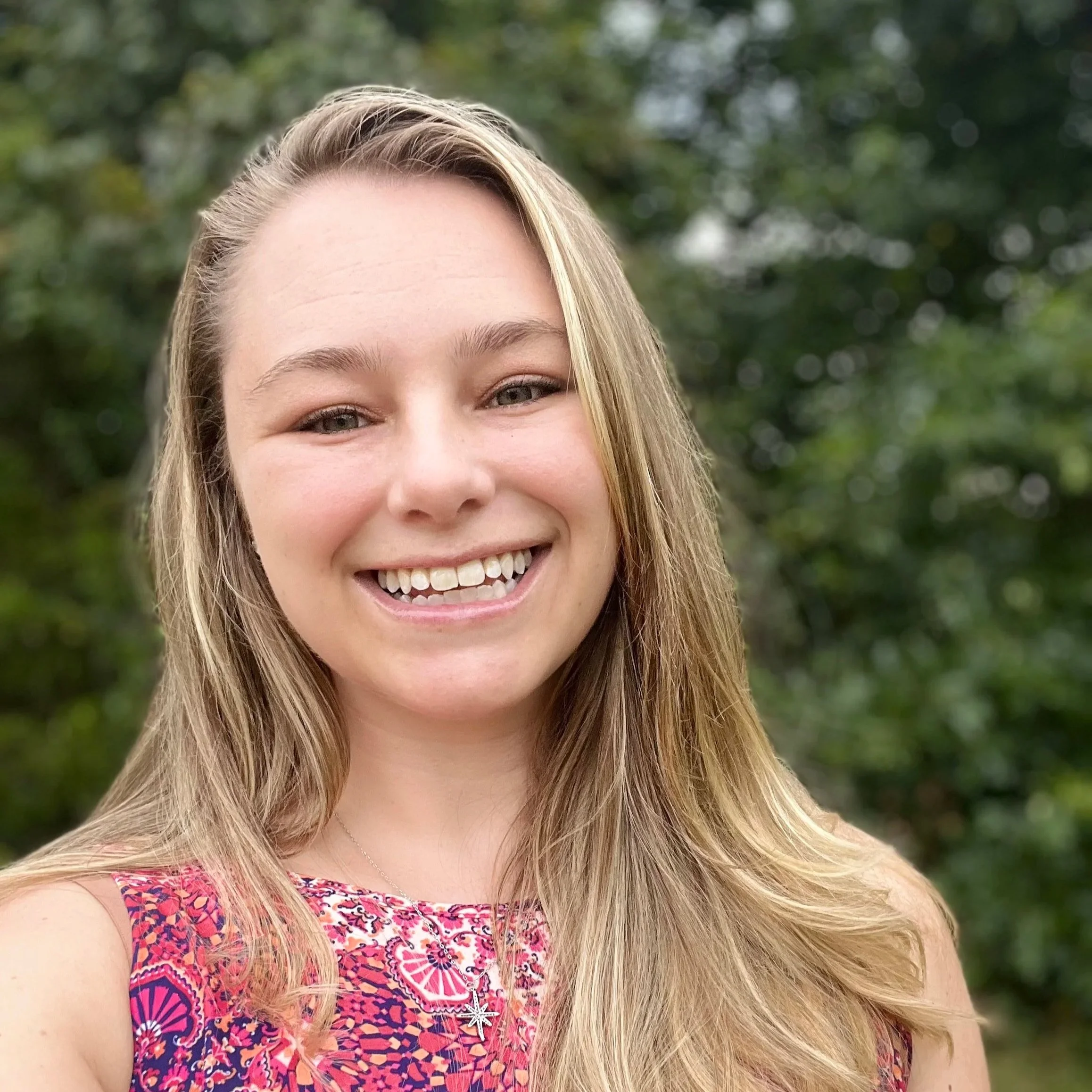 Close-up of a smiling woman with blonde hair, wearing a colorful patterned top, outdoors with blurred green foliage in the background.