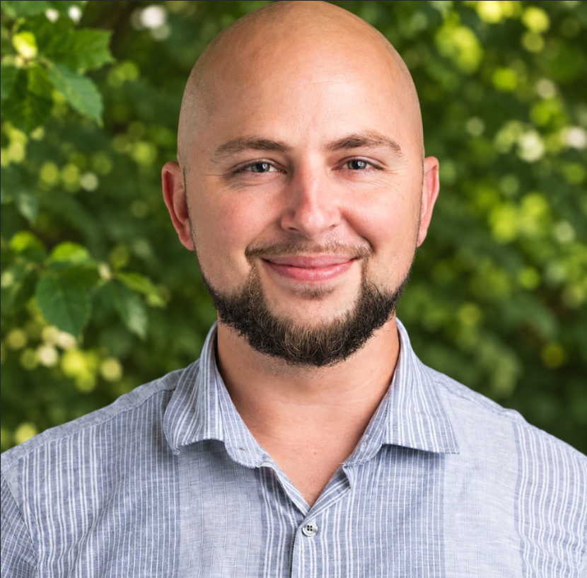 A young man with a bald head and a beard, smiling, wearing a light blue collared shirt, against a plain white background.