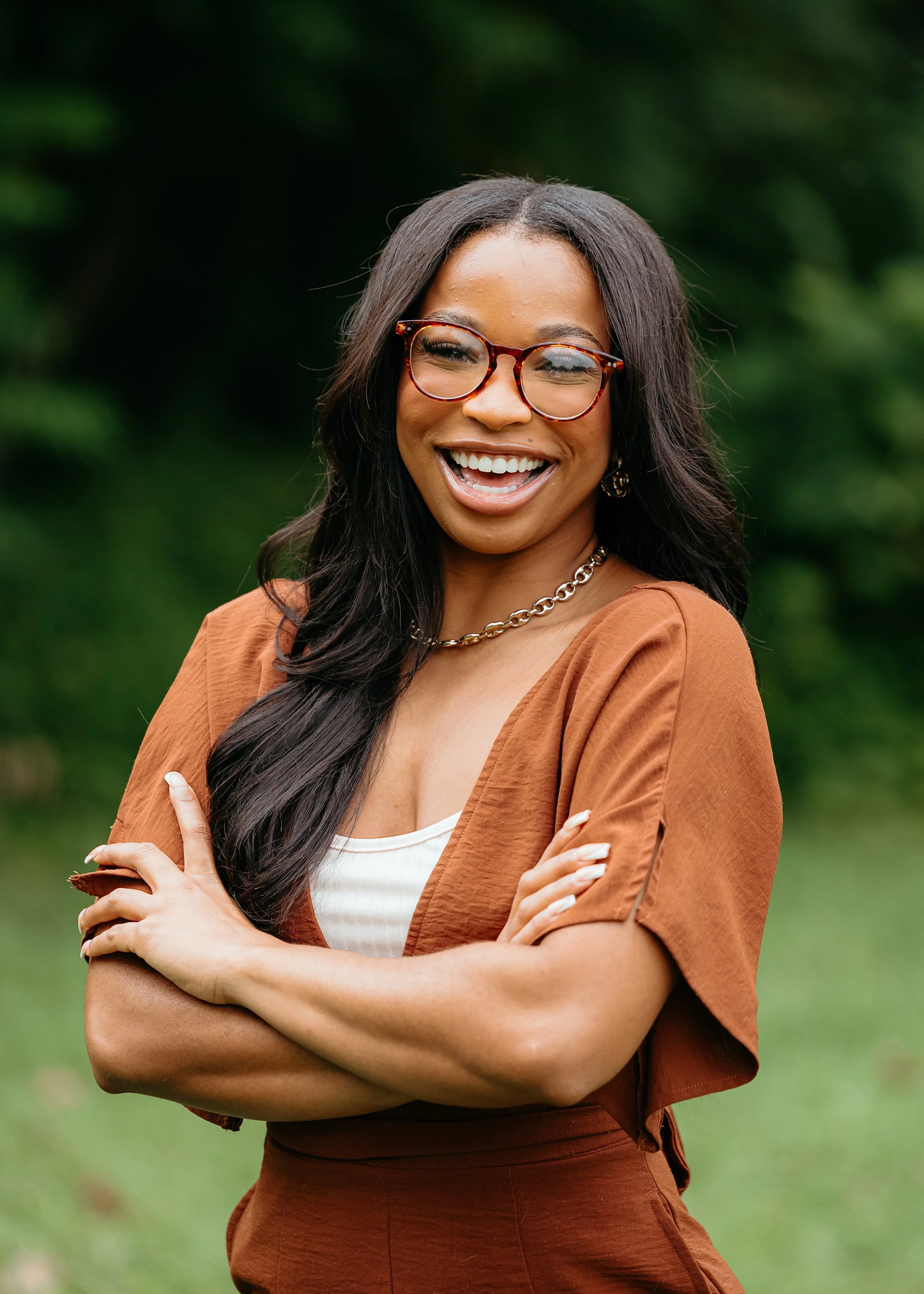 Young woman outdoors laughing, wearing glasses, brown top, and chain necklace, with arms crossed, in front of a blurred green background.