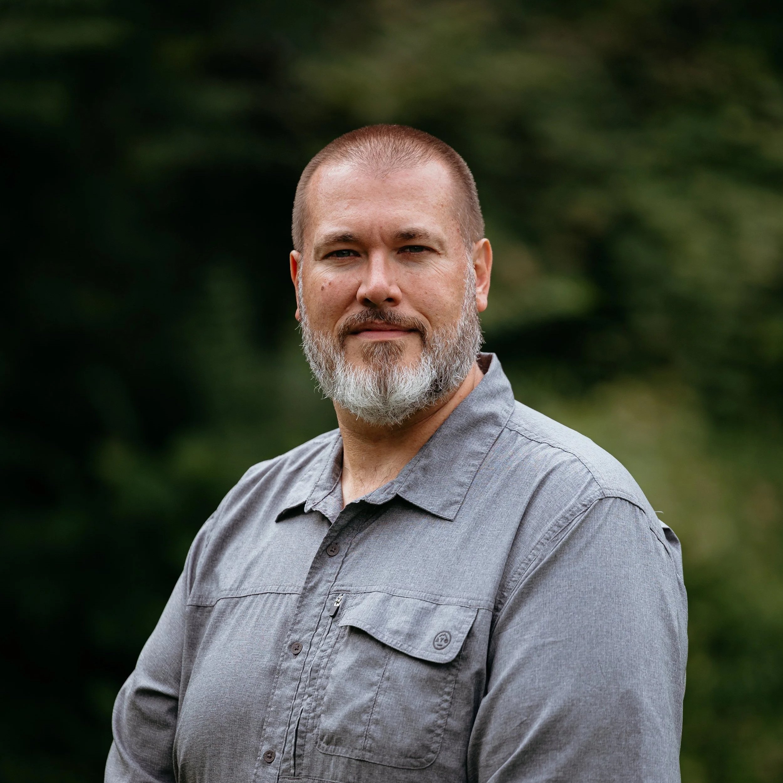 A man with a gray beard and mustache, short hair, and a neutral expression, wearing a gray button-up shirt, standing outdoors with a blurred green background.