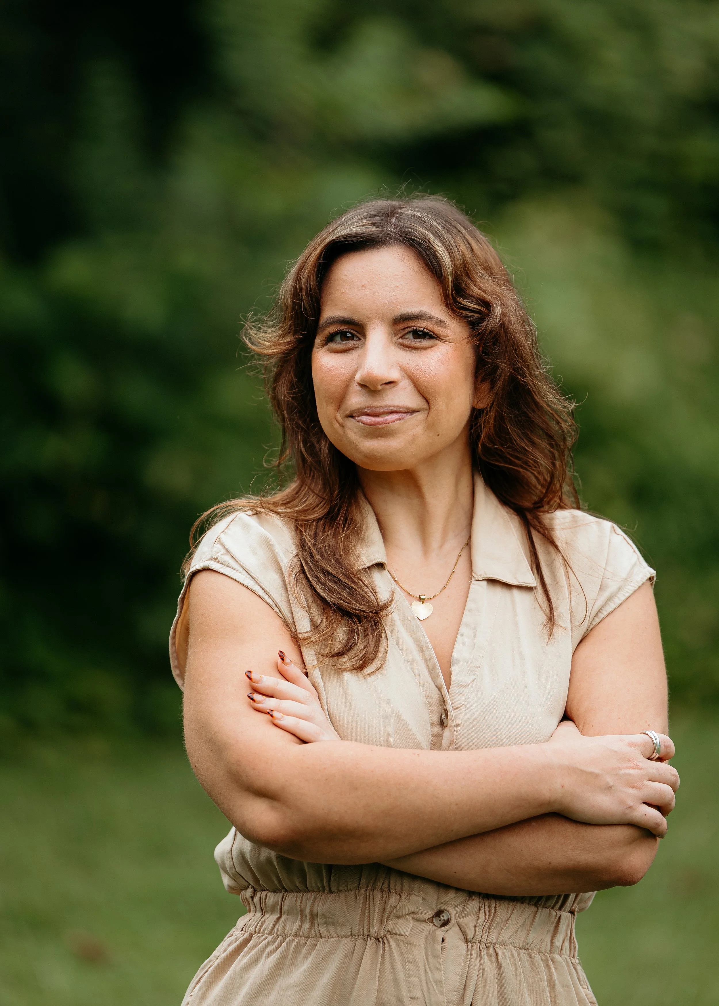 A woman with long, wavy brown hair standing outdoors with her arms crossed, smiling slightly, wearing a beige shirt and beige high-waisted pants, with a blurred green background.