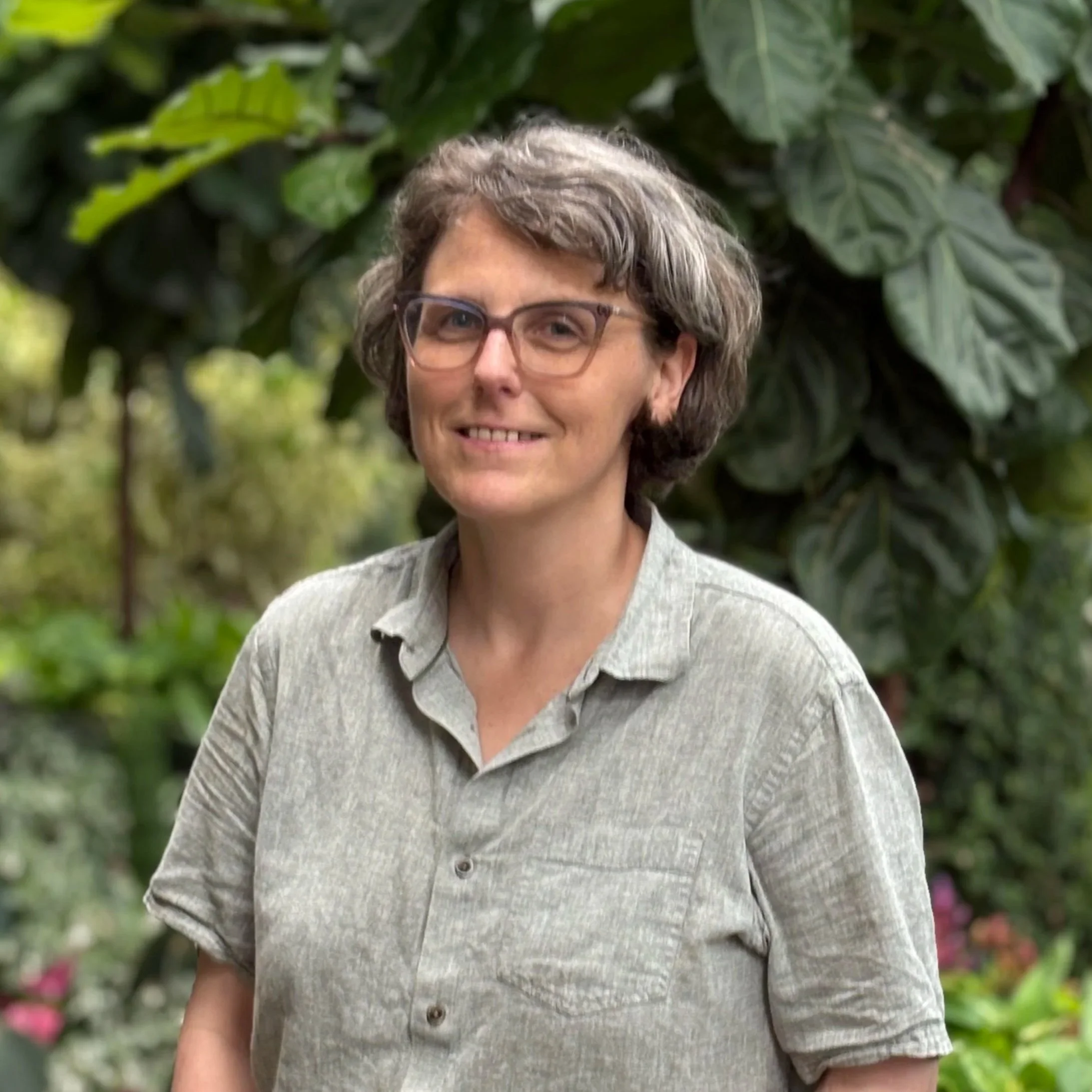 A woman with short gray hair and glasses, wearing a light gray button-up shirt, standing outdoors among green foliage.