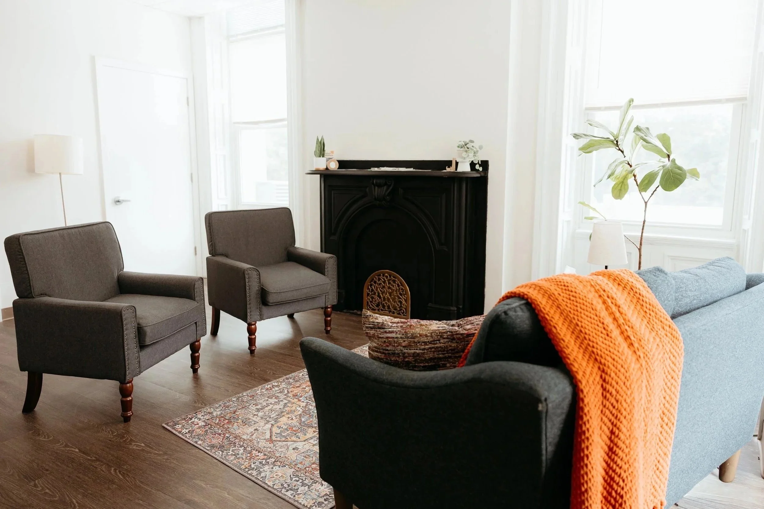 A cozy living room with large windows, two gray armchairs, a black fireplace, a plant in a white pot, a small table with a lamp, and a sofa with an orange blanket.