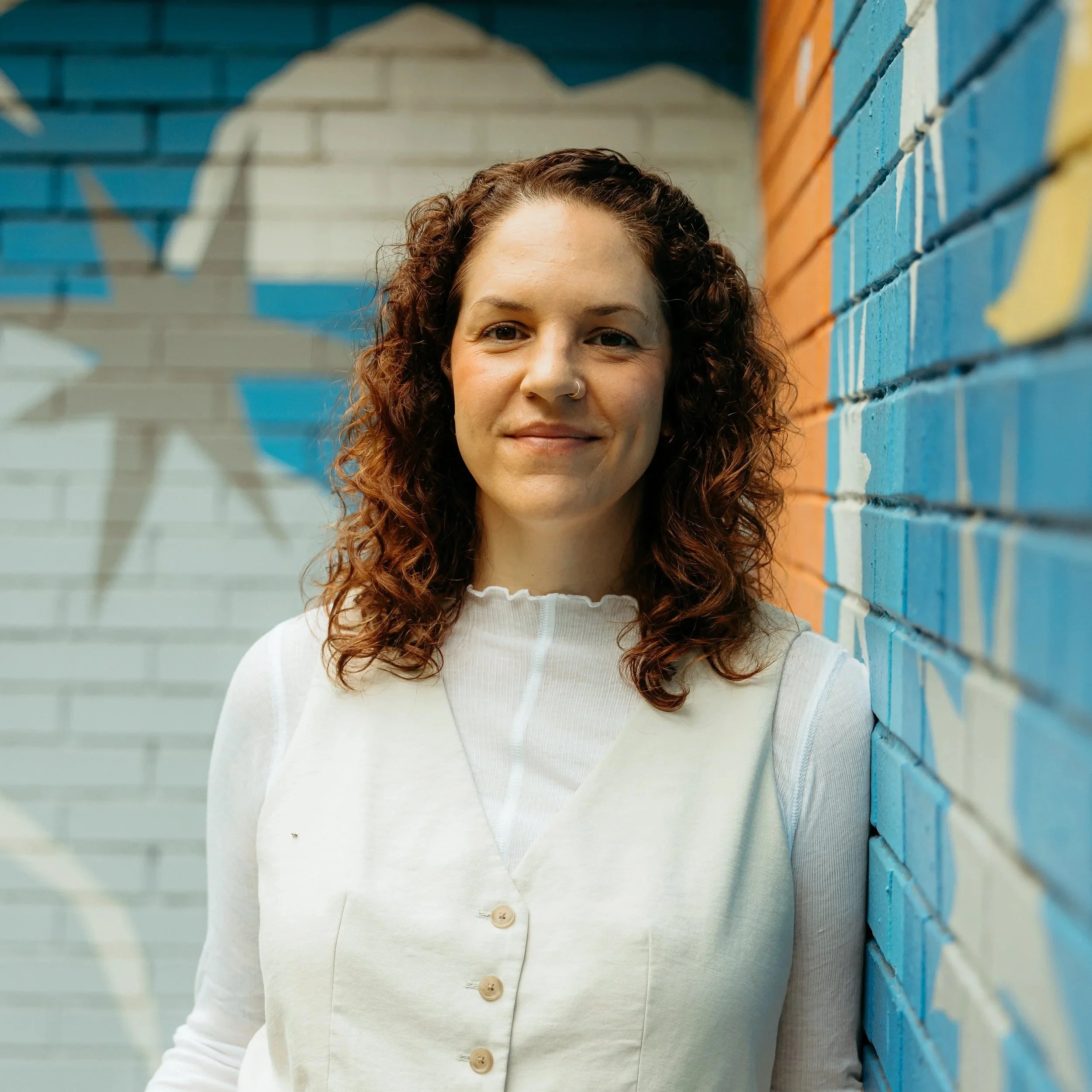 A woman with curly brown hair, smiling, standing next to a colorful brick wall with blue, orange, and white paint.
