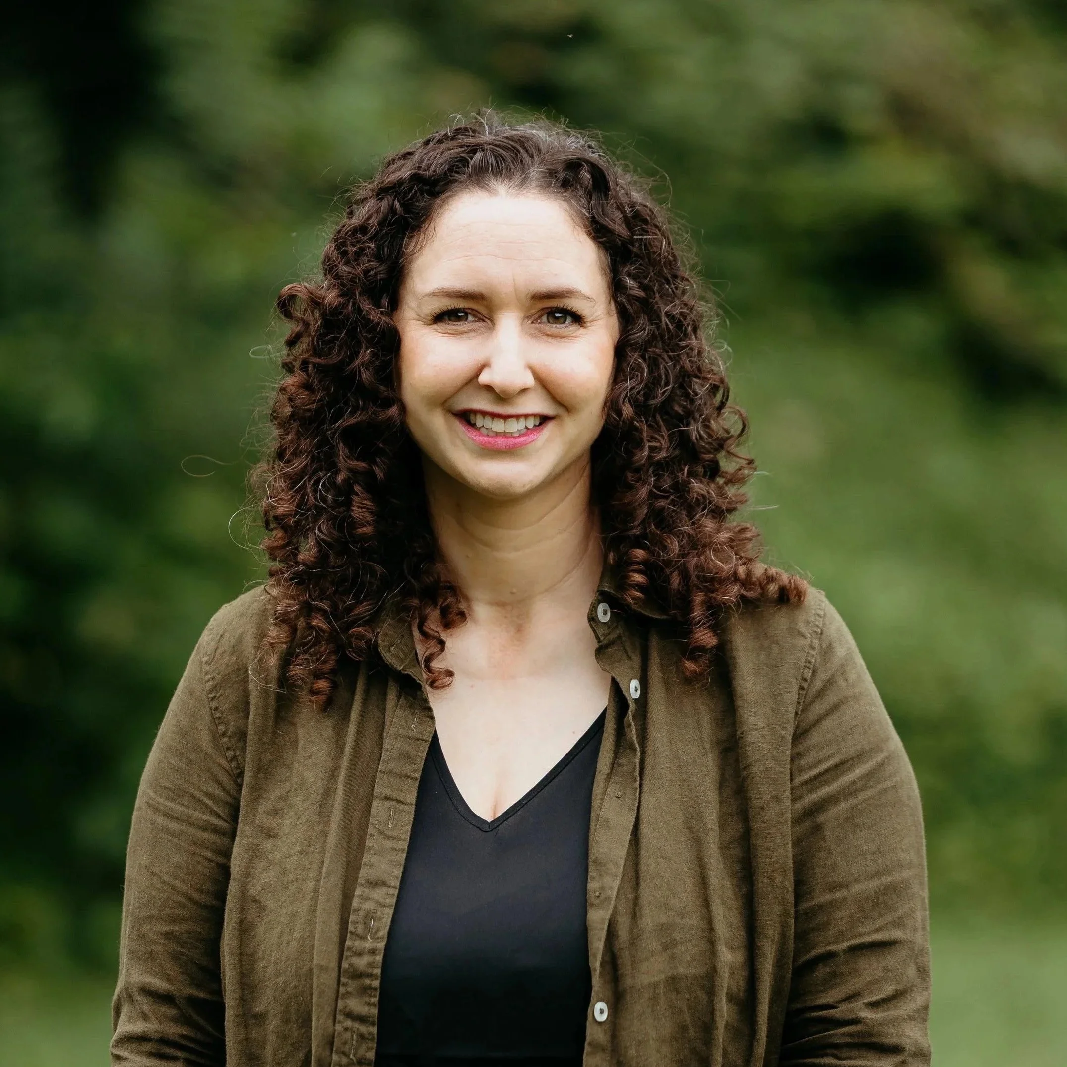 Close-up of a smiling woman with curly brown hair, wearing a brown jacket and a black top, standing outdoors with green foliage in the background.