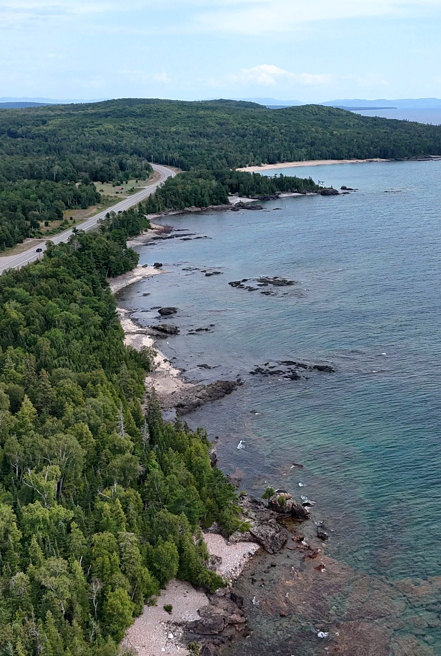 Aerial view of a coastal landscape with a winding road, dense green forest, rocky shoreline, and calm blue ocean waters under a partly cloudy sky.