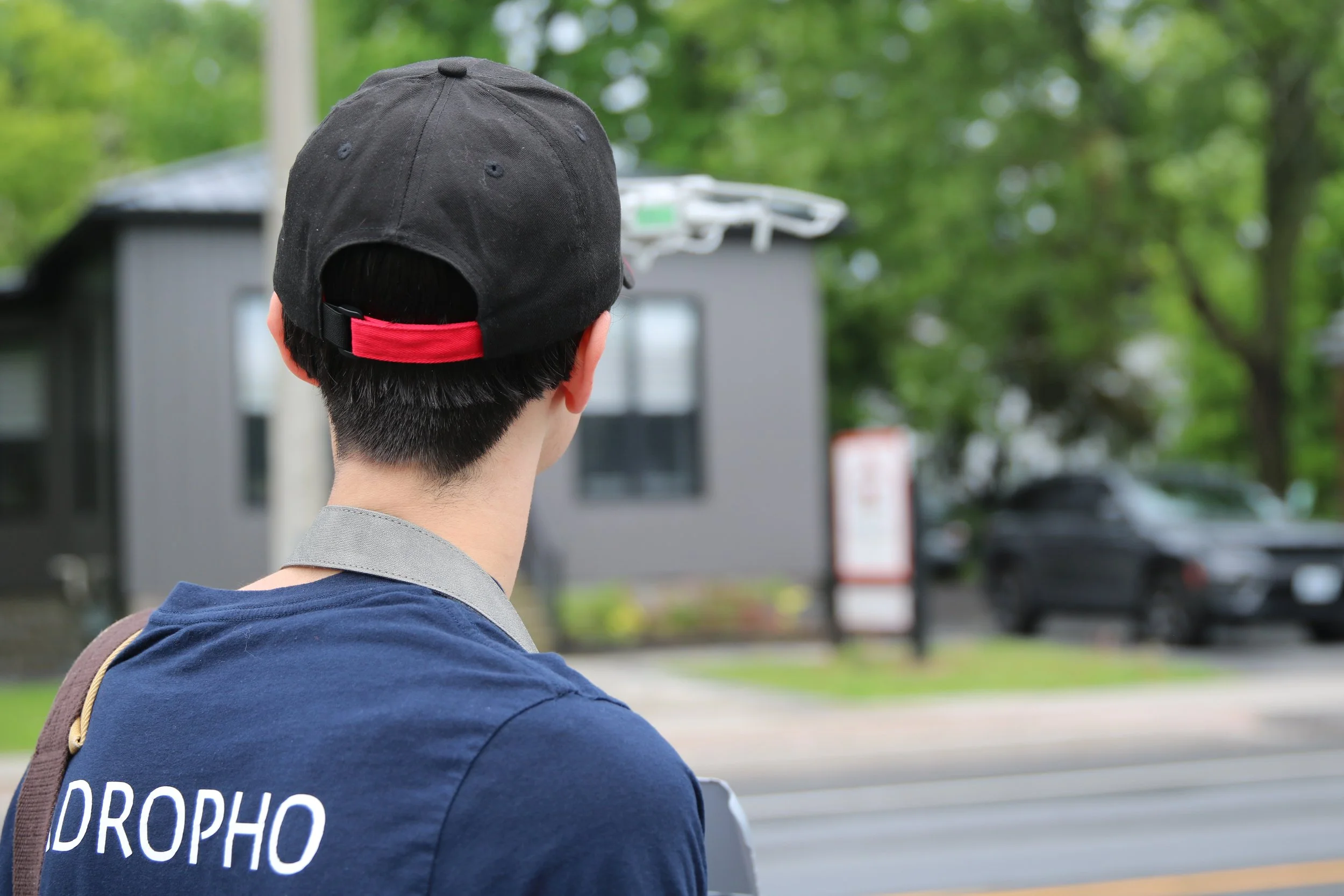 Back view of a person wearing a black cap with a red strap and a navy blue shirt with part of the word 'DROP' visible, standing outdoors with green trees and blurred buildings in the background.
