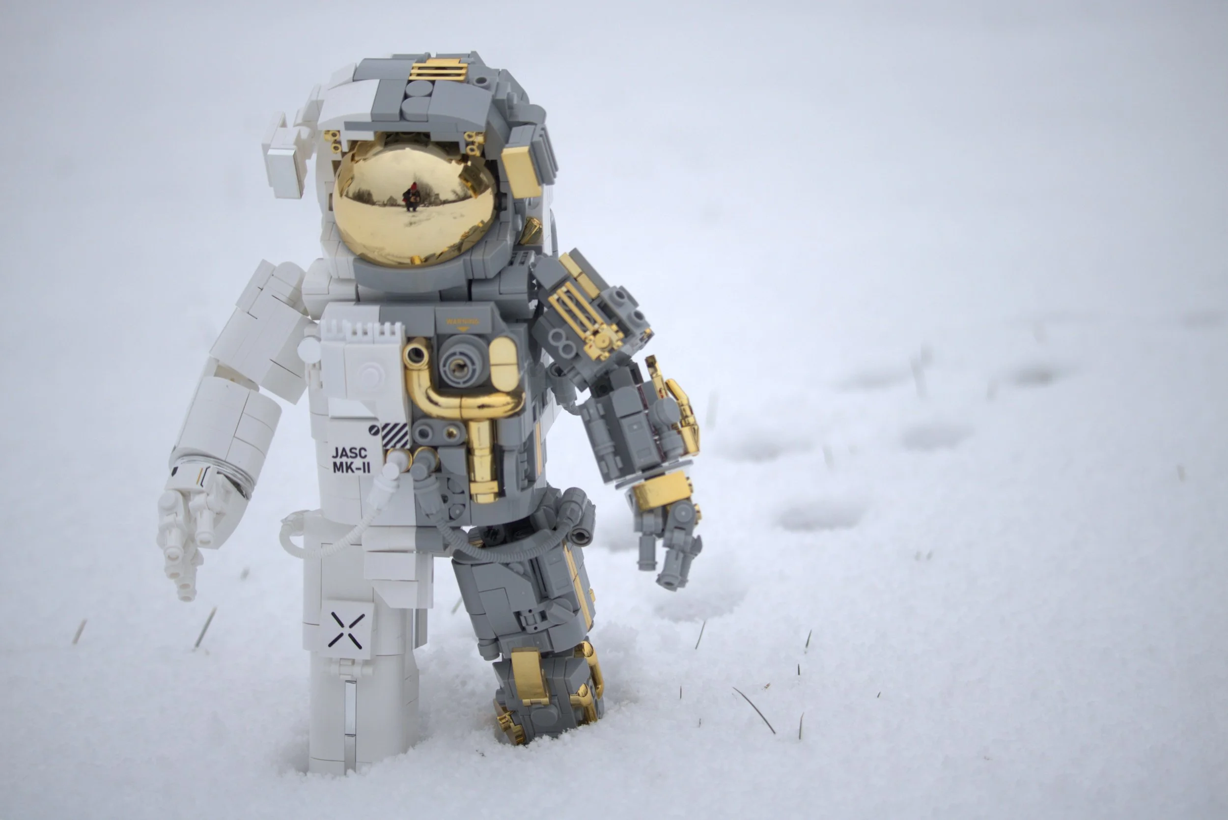 A LEGO astronaut figure standing on snow, with a reflective helmet visor showing a person taking the photo and a snowy landscape in the background.