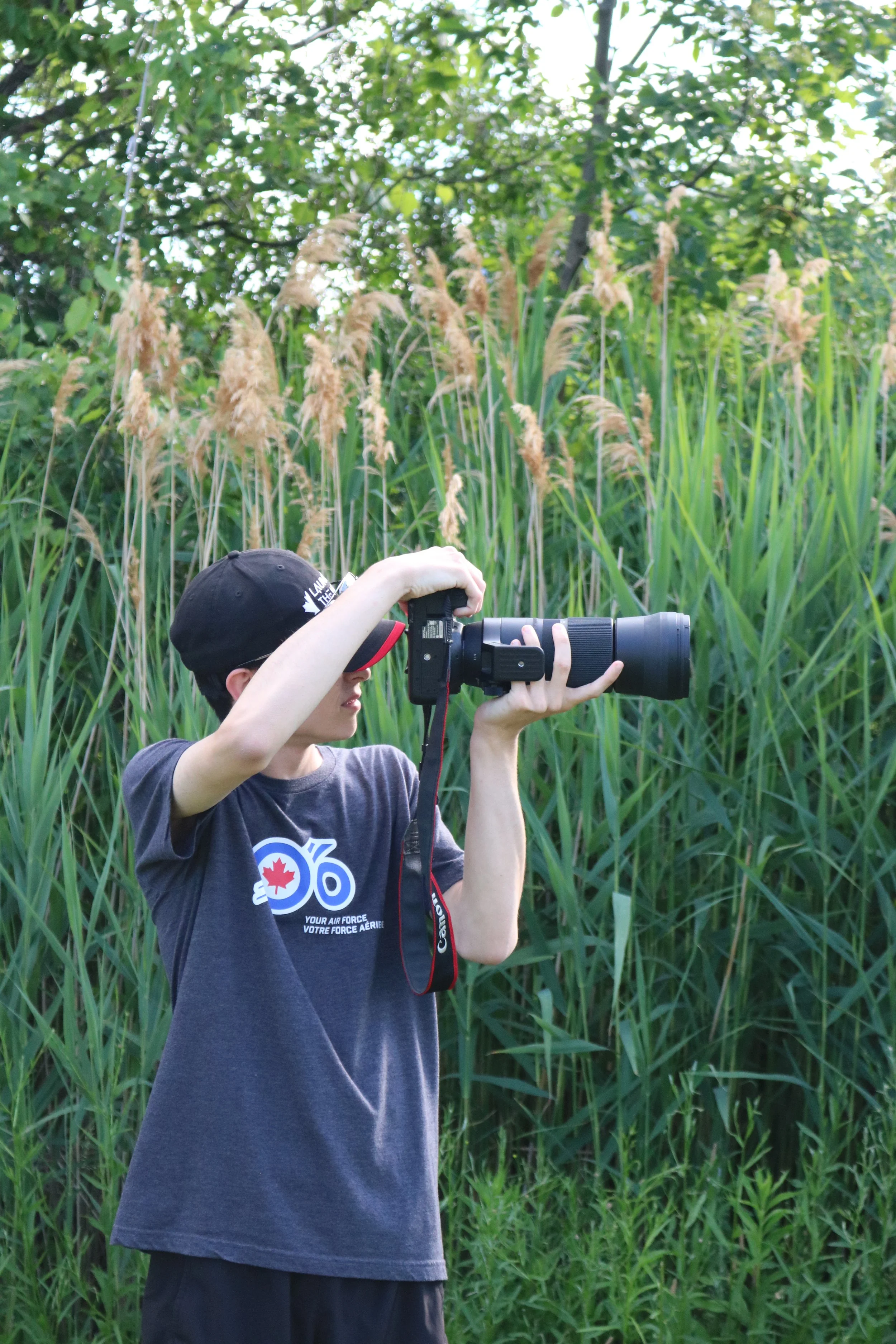 A person wearing a dark T-shirt and a cap, holding a camera with a large lens, standing outdoors in front of tall grass and green leafy trees.