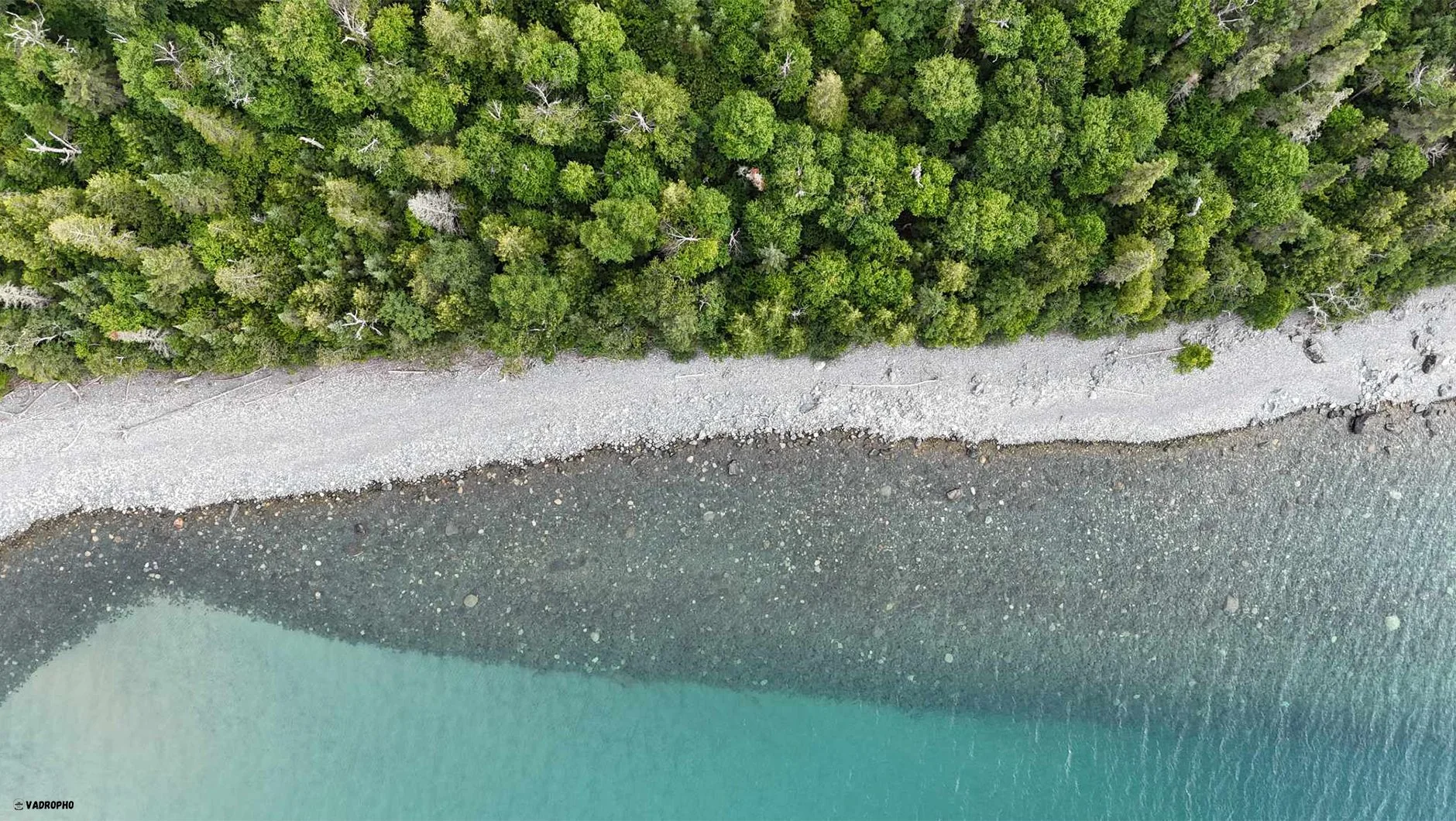 An aerial view of a beach with a gravel shoreline and a dense forest of green trees along the coast, adjacent to a calm body of water.