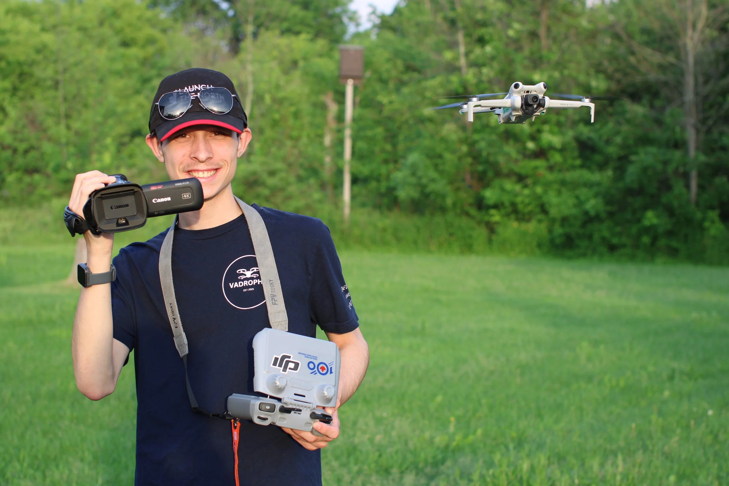A young man holding a remote control for a drone, smiling, outdoors in a grassy area with trees in the background, another drone flying in the air nearby.