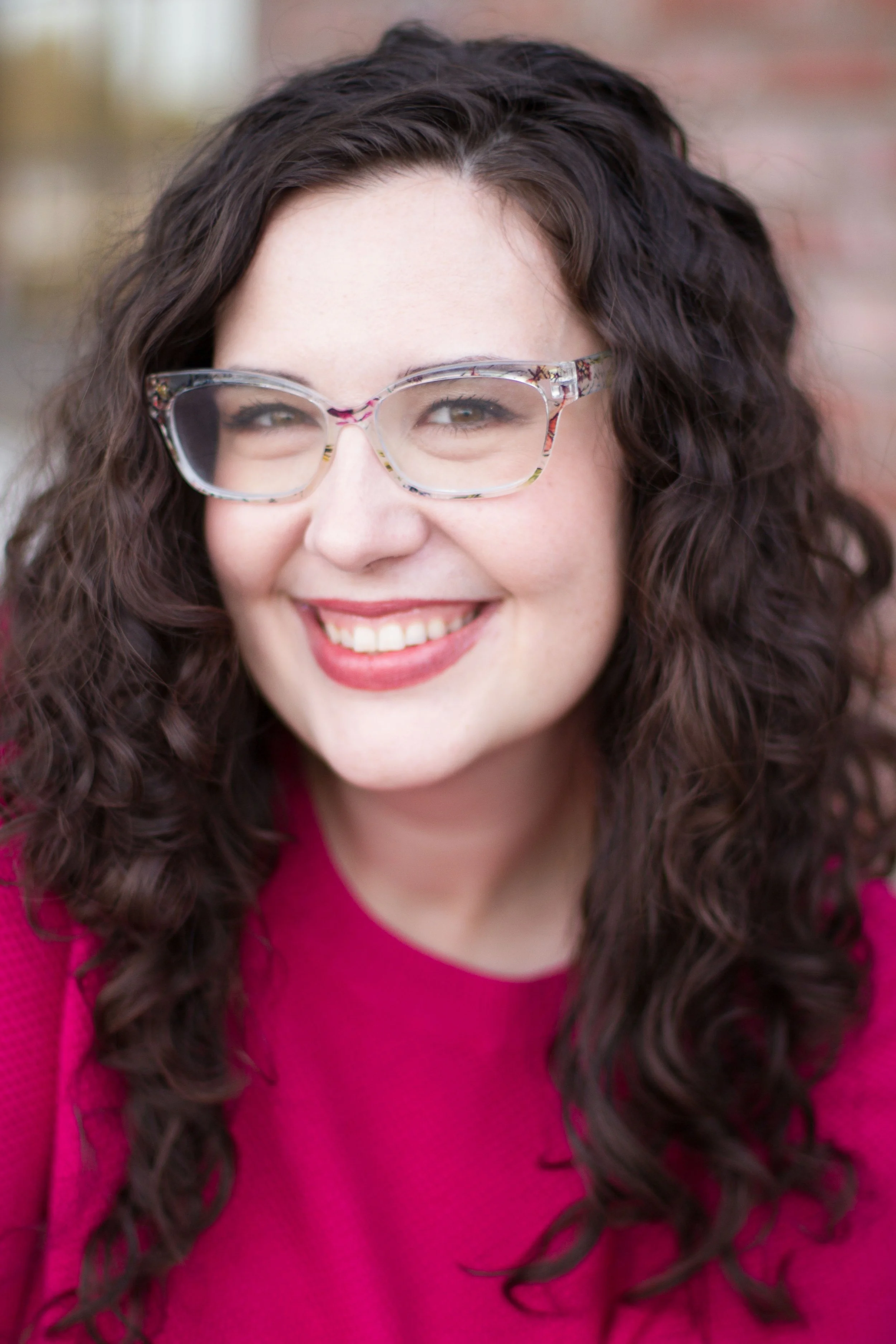 A woman with long, curly dark hair, wearing colorful glasses, smiling, and dressed in a pink top.
