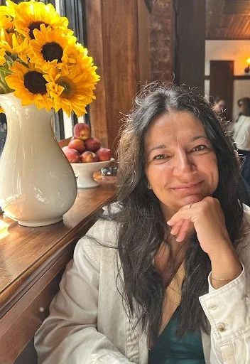 A woman with long dark hair smiling, sitting near a wooden table with a large white vase of sunflowers and a bowl of apples.