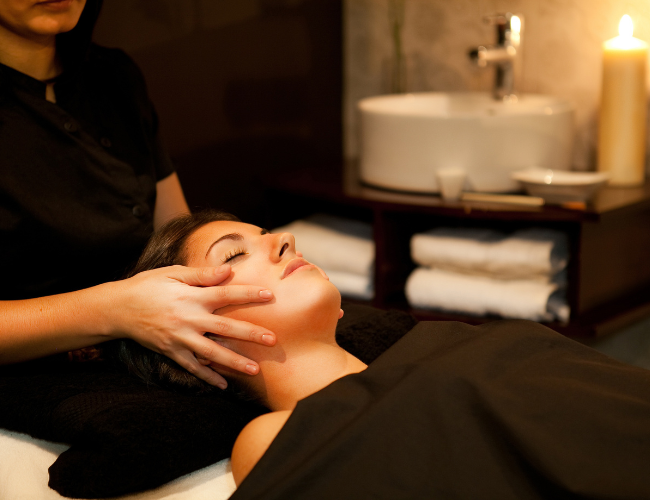 A woman receiving a facial treatment at a spa, lying on a treatment bed with eyes closed, while a therapist massages her face. The spa room features candles, a sink, and towels in the background.