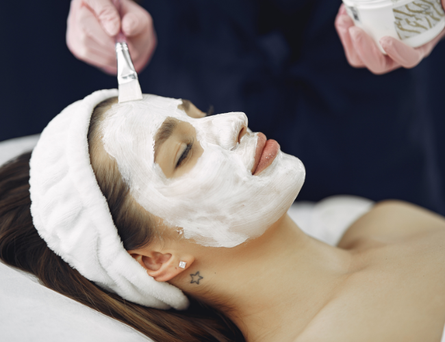 Woman receiving a facial treatment with a white cream mask, lying with a headband, while a skincare professional applies cream with a brush.