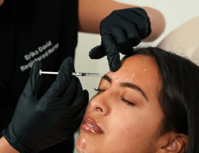 A woman receiving a cosmetic injection in her forehead from a medical professional wearing black gloves