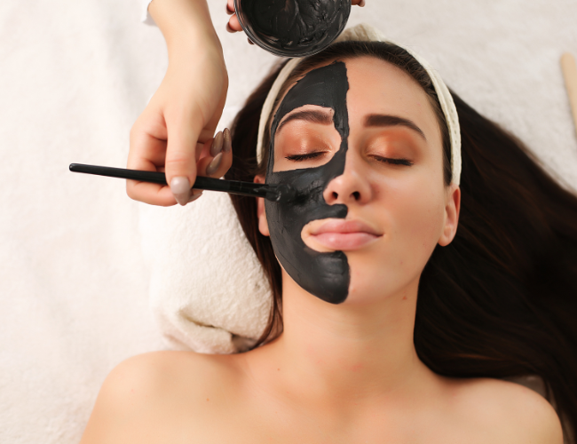 A woman with closed eyes receiving a facial mask treatment, with black mask being applied by a professional using a brush at The Harrison Cincinnati Ohio Day Spa