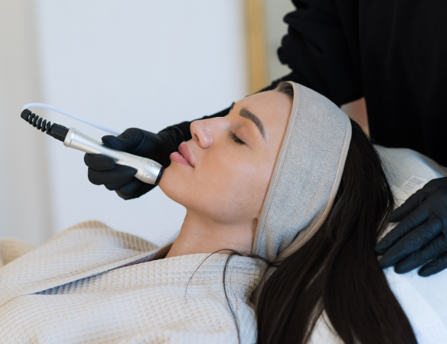 A woman with a gray headband receiving a facial treatment with a handheld device from a technician wearing black gloves