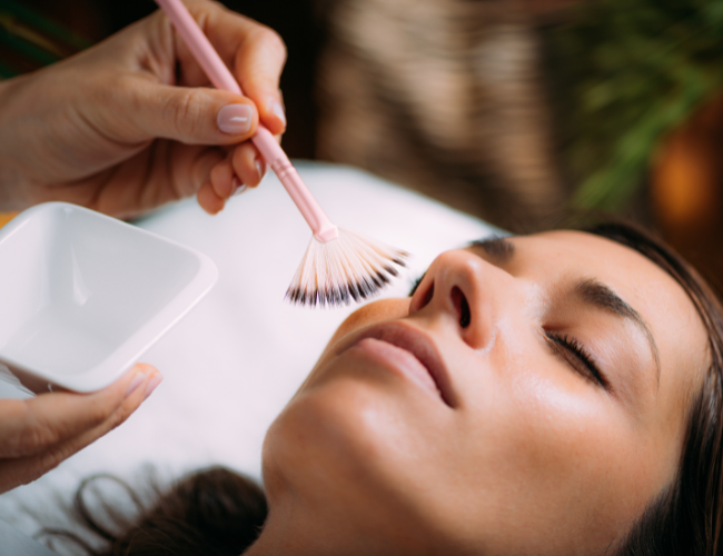 A woman receiving a facial treatment, with a technician using a makeup brush on her face.