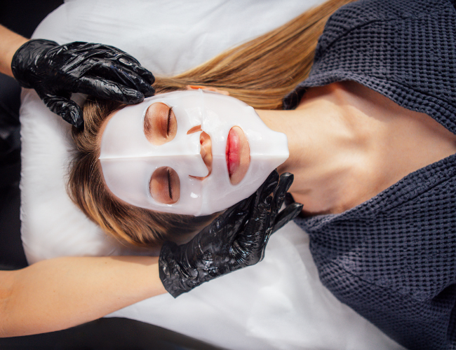A woman receives a facial treatment while lying down, wearing a white facial mask, with eyes closed, and a spa robe. A practitioner wearing black gloves applies the mask.