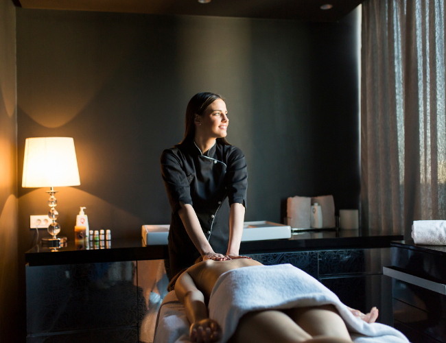 A woman in a dark uniform giving a massage to a client lying face down on a massage table in a dimly lit room with a lit lamp and cabinet in the background.