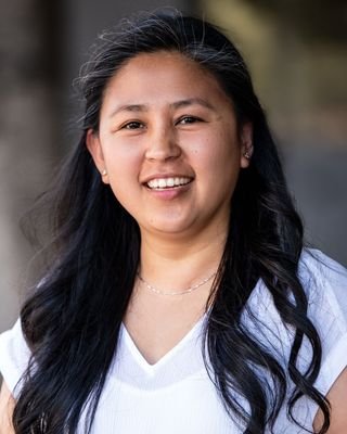 Close-up portrait of a young woman with long black hair smiling outdoors, wearing a white top.