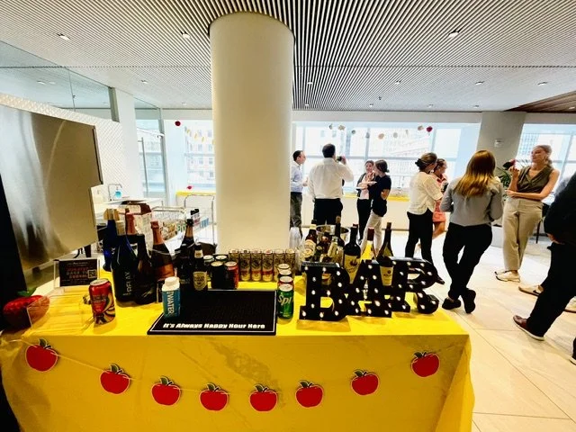 Party table with drinks and snacks, decorated with a yellow tablecloth and red apple banners, in a modern indoor space with people socializing in the background.