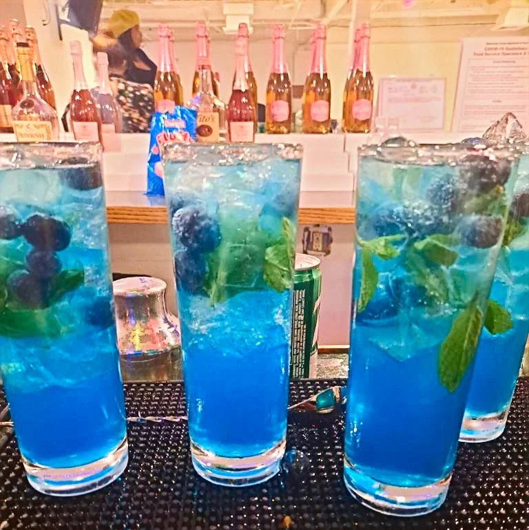 Three tall glasses filled with a bright blue beverage, ice, blueberries, and mint leaves on a bar counter with liquor bottles in the background.