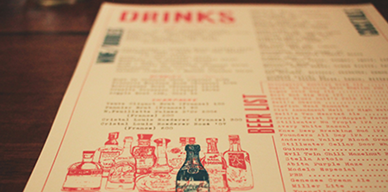 Close-up of a restaurant menu on a wooden table, with a focus on the bottom section showing illustrations of condiment bottles.