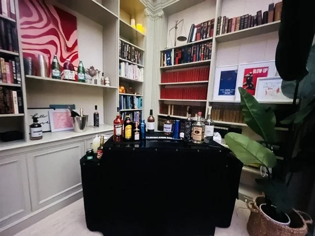 A room with white bookshelves filled with books and decorative items, a black table with various bottles and drinks, and a large green plant in a woven basket.