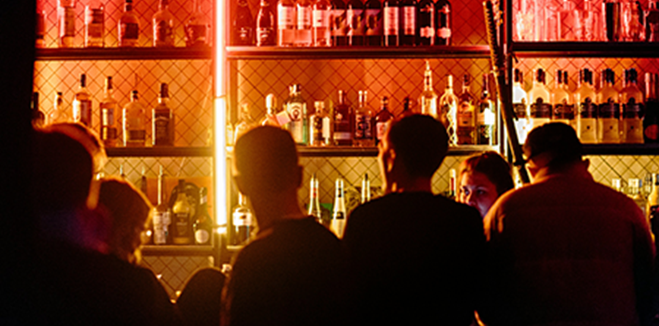 People sitting at a bar with illuminated liquor bottles on shelves behind them, warm orange lighting, and a modern, stylish atmosphere.