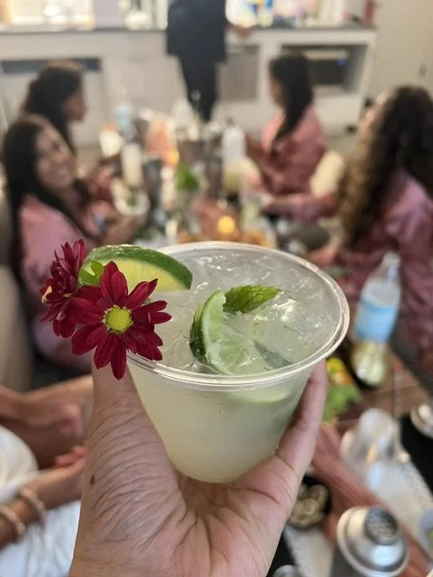 A hand holding a cocktail garnished with lime wedges, mint leaves, and a red flower, with a group of women sitting around a table in the background.
