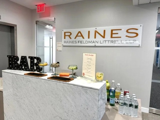 Reception desk with a sign that reads 'RAINES' and 'RAINES FELDMAN LITTRELL LLP'; a large decorated 'BAR' sign; three glass containers with decorative items; menu stand; bottled water and soft drinks on the right side.