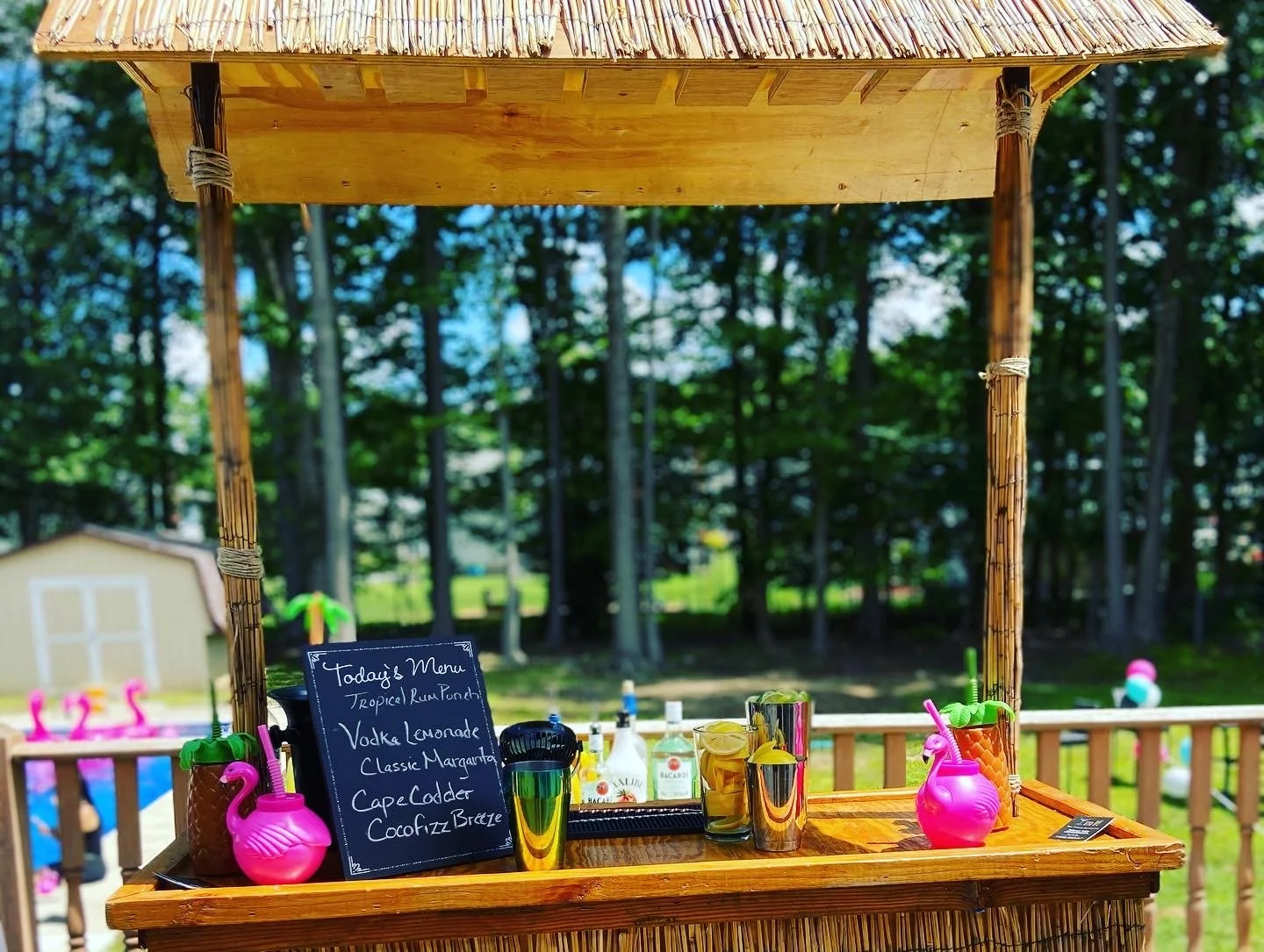 A tiki bar stand made of wood and bamboo with a small blackboard menu listing tropical rum punch, vodka lemonade, classic margarita, cape codder, and cocofizz brite. The stand has colorful plastic flamingo drink holders, metal shaker, and liquor bott
