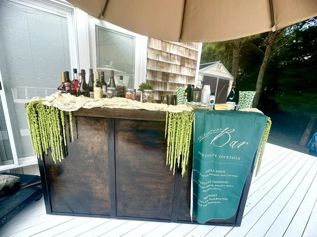 Bar setup on a wooden outdoor table with bottles of alcohol, mixers, and a teal banner advertising specialty cocktails, under a large umbrella.