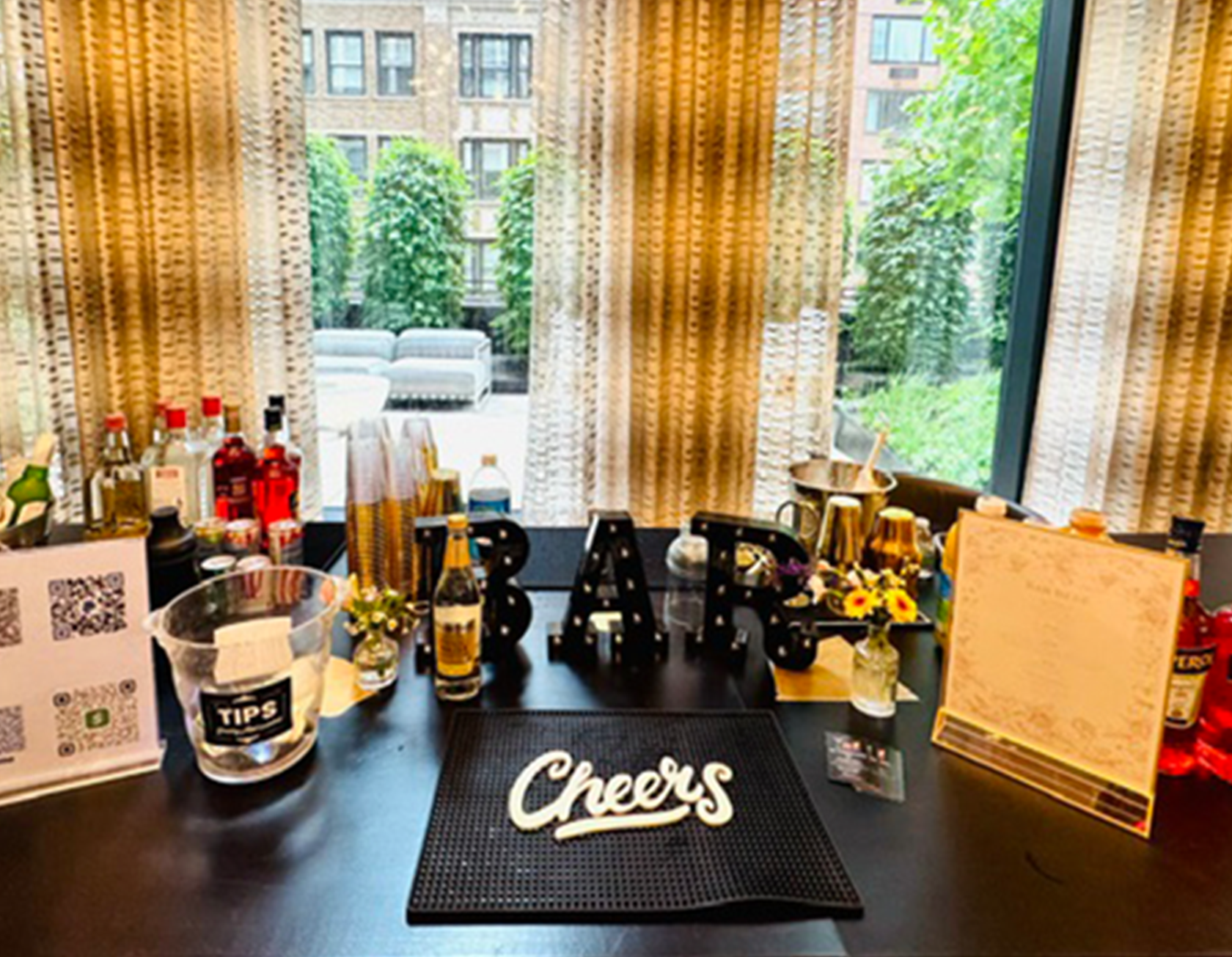 Indoor scene with a table having bottles, flowers, and a sign that says 'Cheers'; behind the table, a large window with sheer curtains revealing an outdoor patio with lounge chairs and green trees.