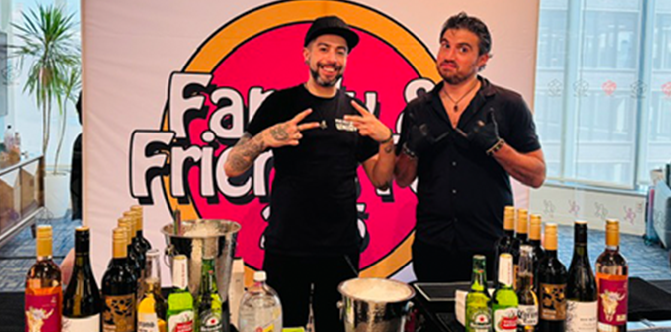 Two men standing at a table with numerous bottles of alcohol in front of them, smiling and making peace signs. Behind them is a colorful backdrop with text that says 'Fry' and 'Friender.'