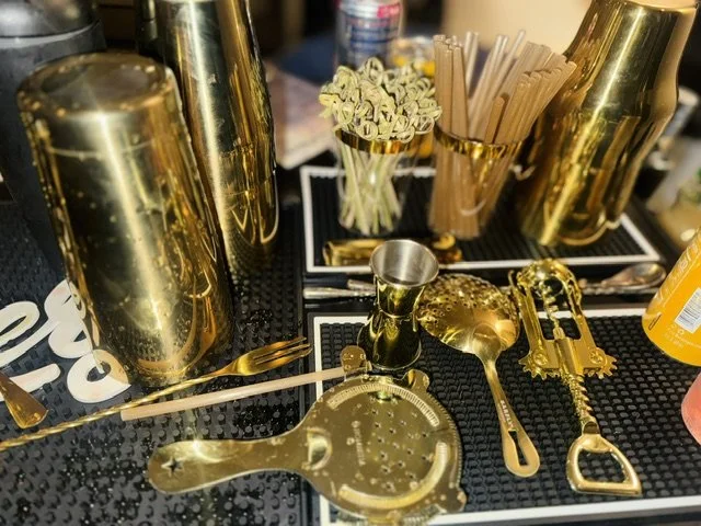 Bar_counter with gold barware and utensils, including a cocktail shaker, strainer, spoon, jigger, knife, and straw holder, all placed on a black textured surface.
