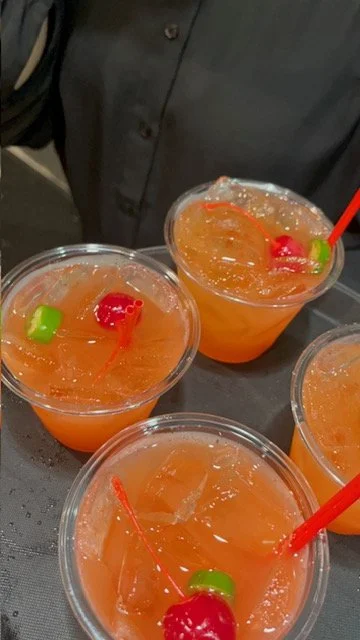 Three plastic cups with orange-colored beverages, garnished with cherries and green candy, and red straws on a black surface.