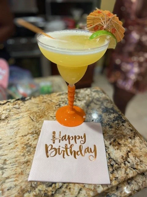 A yellow cocktail with a lime wedge and small paper umbrella in a decorated glass on a birthday table with a "Happy Birthday" napkin.