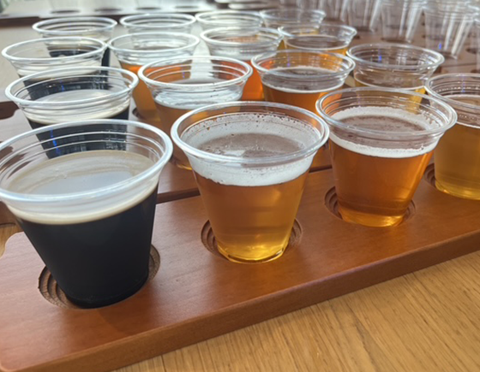 A wooden tasting tray holding multiple plastic cups filled with different shades of beer, placed on a wooden table.